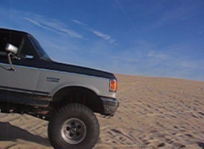 1989 Ford Bronco - A two-tone 1989 Ford Bronco navigates sandy terrain under a clear blue sky.