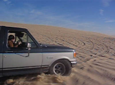 1989 Ford Bronco - A two-tone Ford Bronco kicks up sand as it drives up a dune under a clear blue sky.