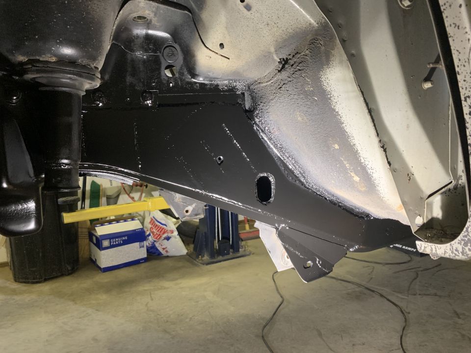 Underside of a vehicle frame coated in black protective paint, showing suspension components.