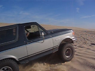 1989 Ford Bronco - A two-tone Ford Bronco kicks up sand as it drives across a desert landscape under a clear blue sky.