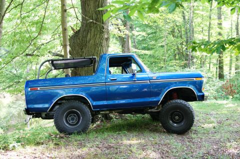 1979 Ford Bronco - A blue Ford Bronco sits off-road in a forest, its lifted suspension and large tires visible.