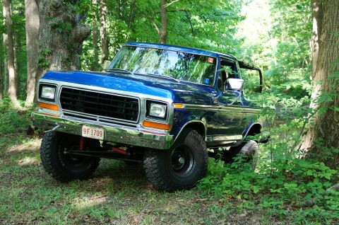 1979 Ford Bronco - This blue 1979 Ford Bronco is parked off-road in a wooded area, showing its front 3/4 profile.