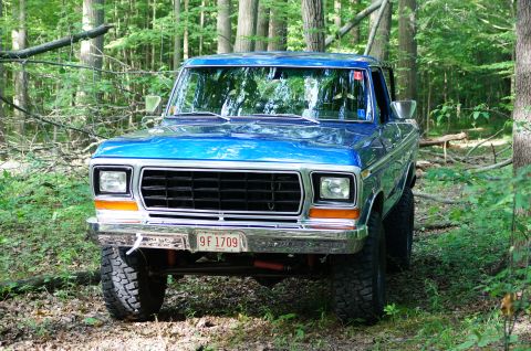 1979 Ford Bronco - A blue 1979 Ford Bronco is parked in a wooded area, showing its front grille and headlights.