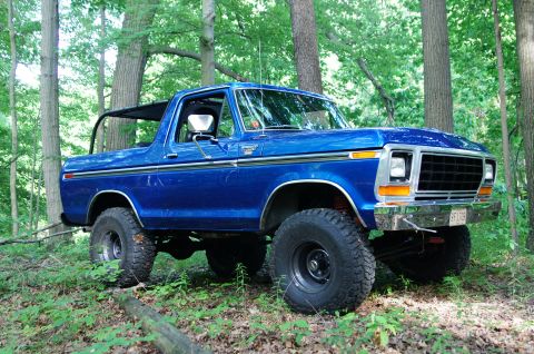 1979 Ford Bronco - A blue Ford Bronco sits off-road in a wooded area, its tires elevated on the uneven terrain.