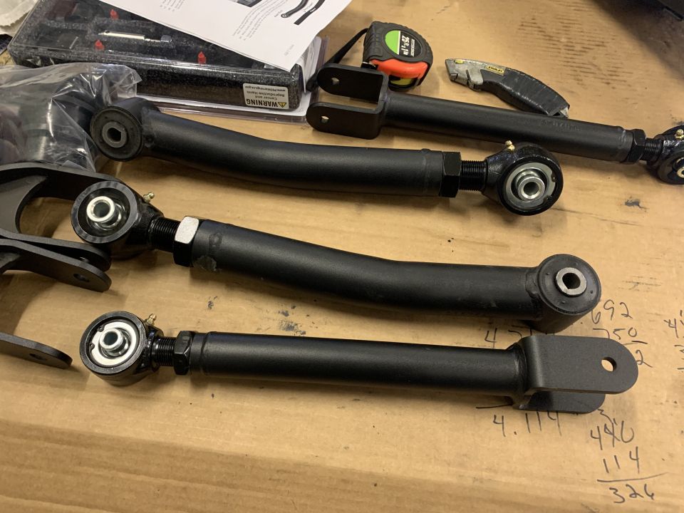 Black adjustable suspension control arms laid out on a wooden surface with a tape measure and utility knife nearby