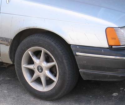 1990 Ford Taurus - Close-up of a car's front wheel and fender area, showing the tire and part of the bumper.