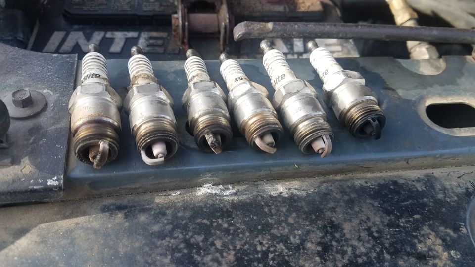 Six used spark plugs lined up on a car hood. Some have black deposits on the tips