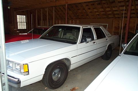 1991 Ford Crown Victoria LTD - A white sedan is parked inside a garage, with another white vehicle partially visible in the foreground and a red car in the background.