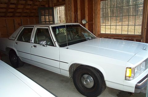 1991 Ford Crown Victoria LTD - A white 1991 Ford Crown Victoria LTD sedan is parked inside a wooden garage, with windows showing trees outside.