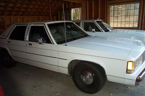 1991 Ford Crown Victoria LTD - Two white sedans are parked side-by-side inside a garage with wooden walls and a large window.