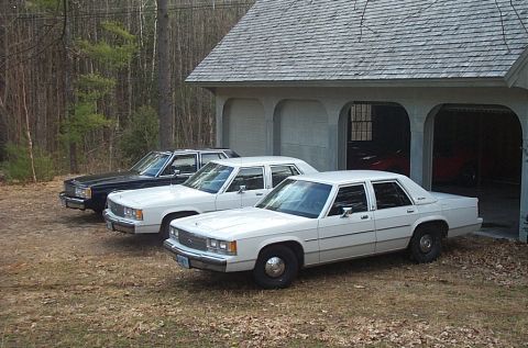1991 Ford LTD Crown Victoria - Three Ford Crown Victoria sedans are parked outside a garage, two white and one black.