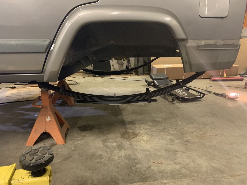 Gray 2000 Jeep Cherokee rear leaf springs being replaced, supported by jack stands
