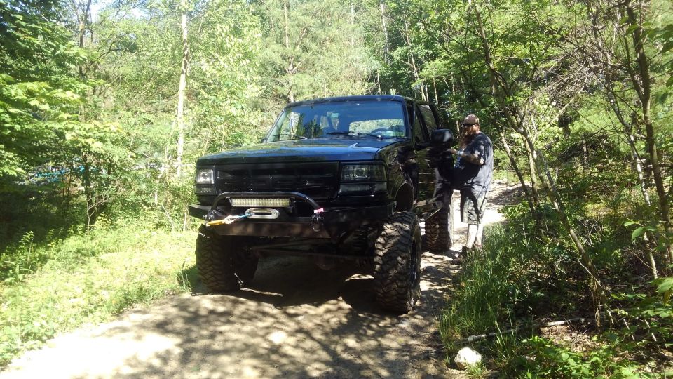 Black lifted Ford Bronco parked on a dirt path in a wooded area with a man standing next to it.