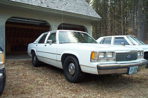1991 Ford LTD Crown Victoria - A white sedan is parked in front of a garage with another vehicle visible behind it.