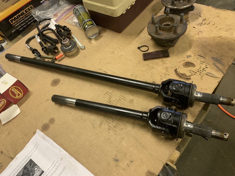 Two black CV axles with U-joints and other automotive parts on a cardboard surface.