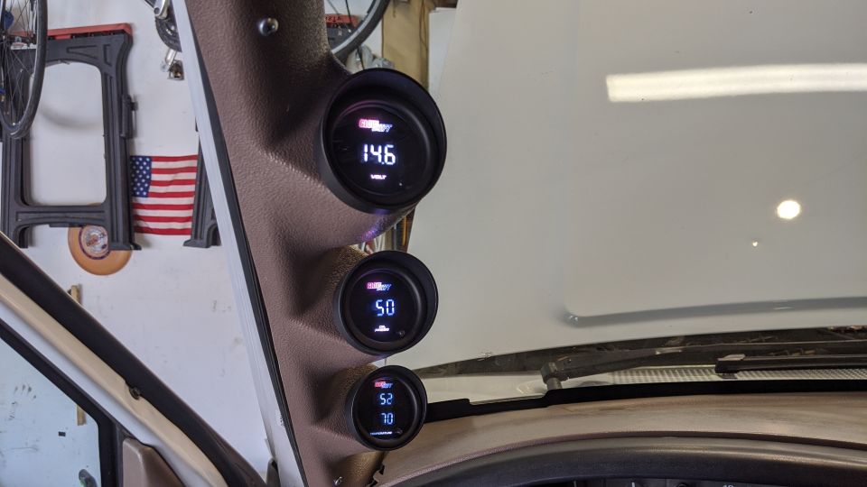 Three digital gauges mounted on a pillar inside a vehicle, showing voltage, temperature, and another reading