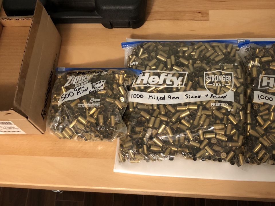 Three plastic bags filled with spent 9mm ammunition casings on a wooden surface. One bag is labeled '500 9mm Mixed Sized + Primed', and two bags are labeled '1000 Mixed 9mm Sized + Primed'. A cardboard box is to the left