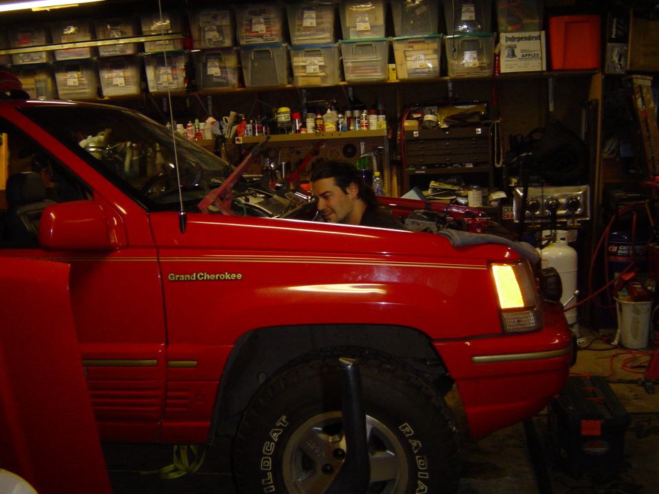 Red Jeep Grand Cherokee in cluttered home garage, man working at the rear, parts bins lining the wall