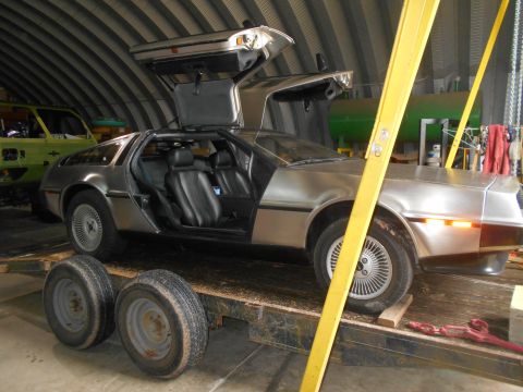 1992 Ford Bronco - A silver DeLorean DMC-12 with its iconic gullwing doors open sits on a trailer, with another vehicle in the background.