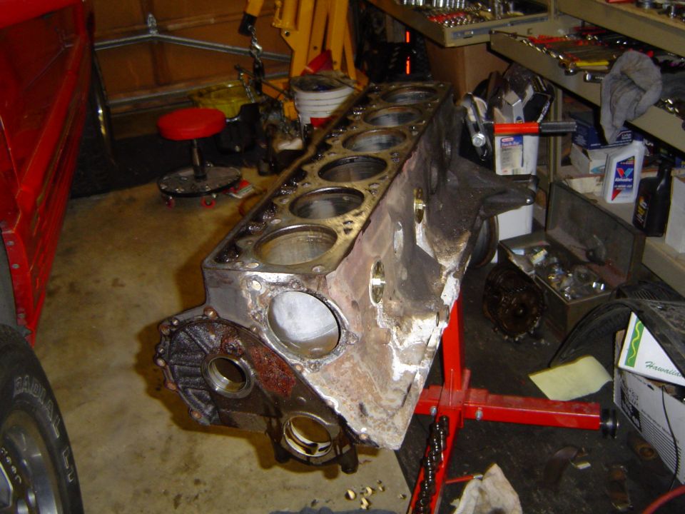Disassembled V8 engine block on garage floor, cylinder bores exposed, one piston in place, red engine stand nearby