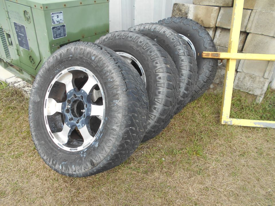 Four off-road tires with silver and black rims on grass, next to a green generator and concrete blocks
