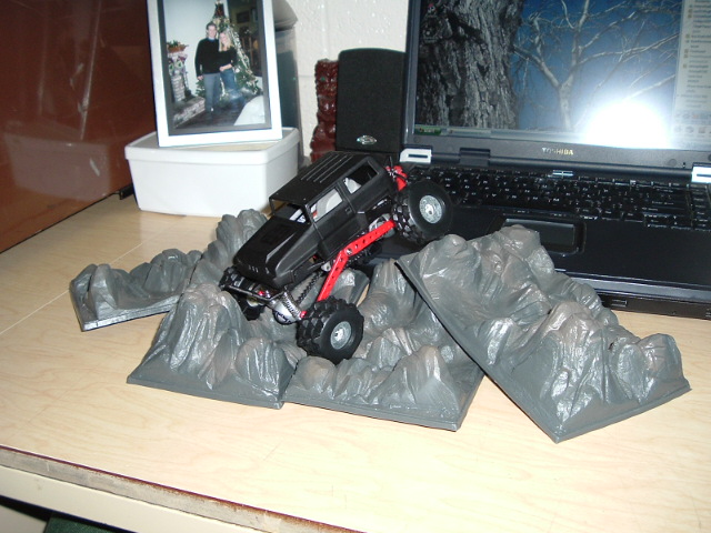 Black toy rock-crawler truck posed on gray plastic rock scenery on a desk beside a laptop