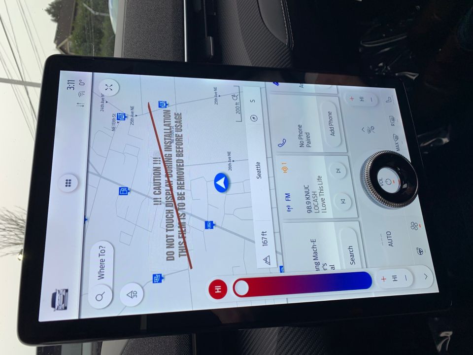Car interior showing a large touchscreen display with navigation map and climate controls. A warning sticker is visible on the screen