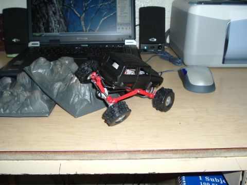 1989 Ford Bronco - This black remote control rock crawler toy features red suspension components and is positioned on a desk next to a laptop and mouse.
