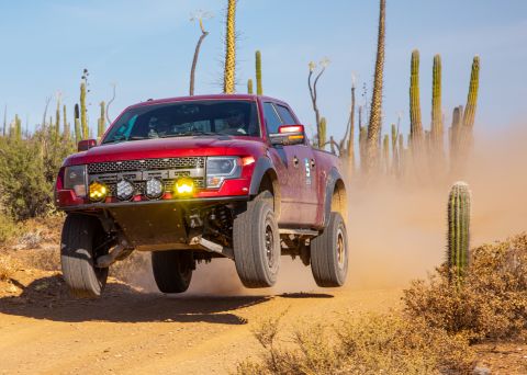 2014 Ford F-150 - Off-road truck airborne on a dirt track, kicking up dust in a desert landscape.
