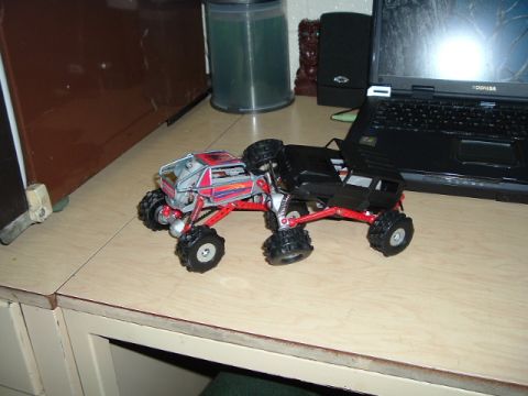 1989 Ford Bronco - Two RC monster trucks are positioned on a desk, one behind the other, with a laptop visible in the background.