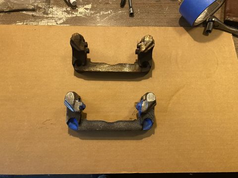 Two brake caliper brackets on a brown surface, one with blue tape