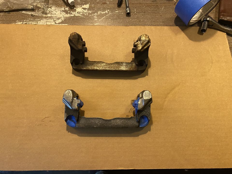 Two brake caliper brackets on a brown surface, one with blue tape