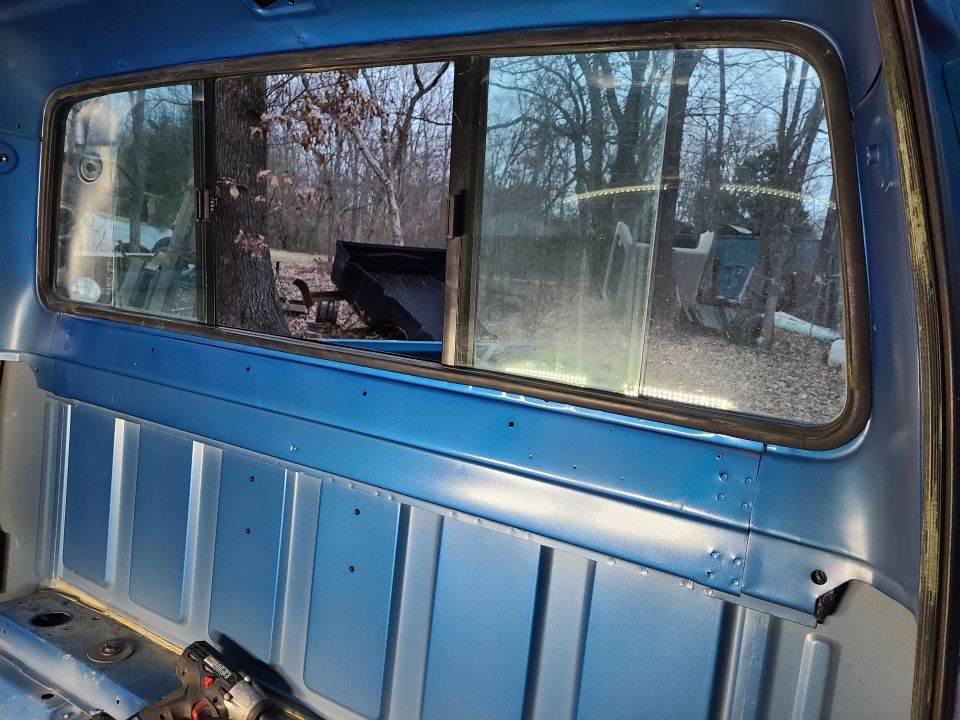 Inside of a blue truck cab, looking out the rear window at trees and a black truck bed