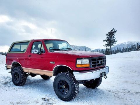 1994 Ford Bronco - A red and tan Ford Bronco sits in a snowy landscape, its lifted suspension and large tires ready for winter conditions.