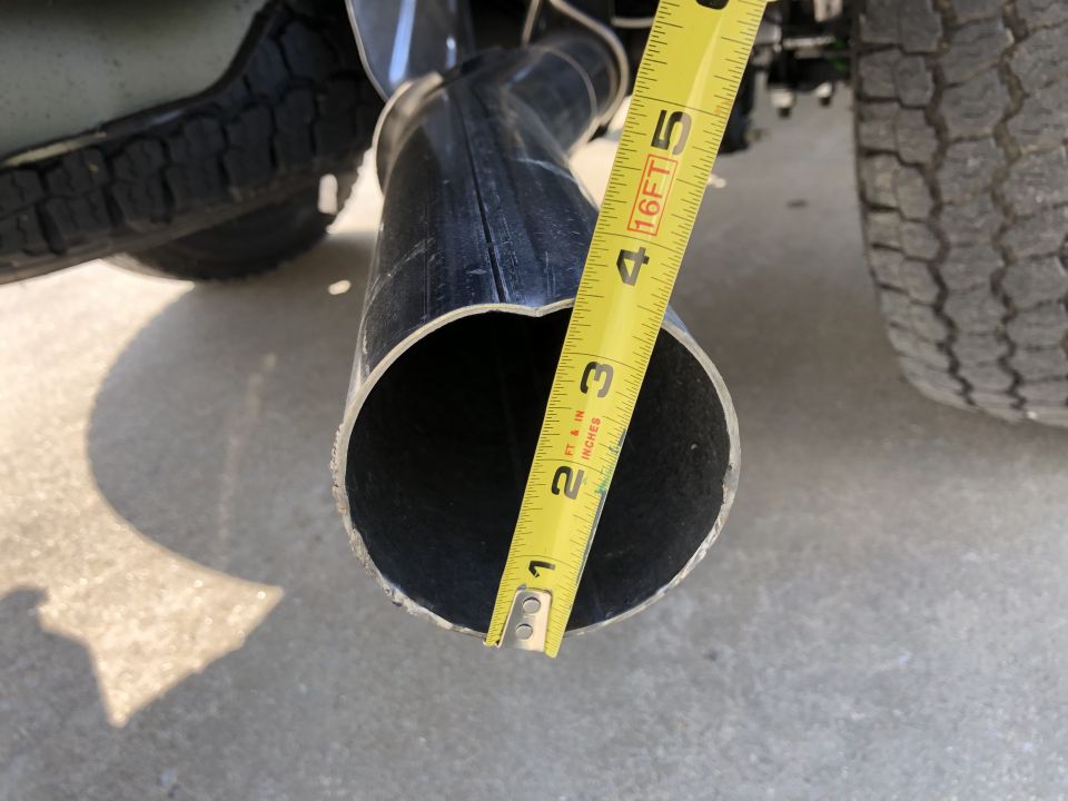 Measuring the diameter of a black exhaust pipe with a yellow tape measure. Tire and concrete visible