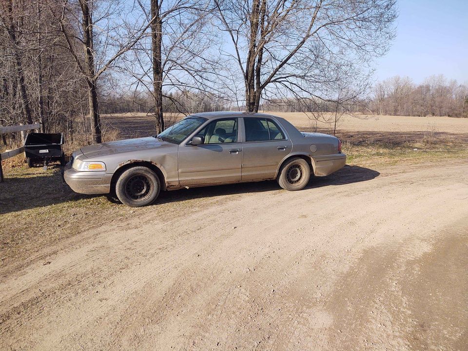 2005 Ford Crown Victoria parked on a dirt road, side view. Car has steel wheels and some rust
