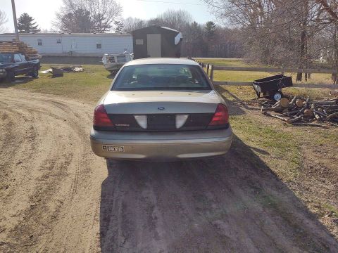 2005 Ford Crown Victoria - A tan Ford Crown Victoria is parked on a dirt road in a rural setting with a trailer and woodpile nearby.