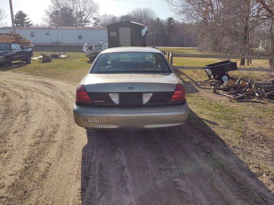 Rear view of a tan Ford Crown Victoria, possibly from the early 2000s, parked on a dirt road