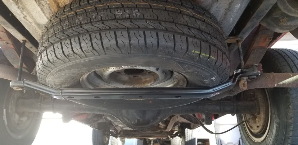 Spare tire and black carrier bar under a truck's rear frame, with rear axle and driveshaft visible