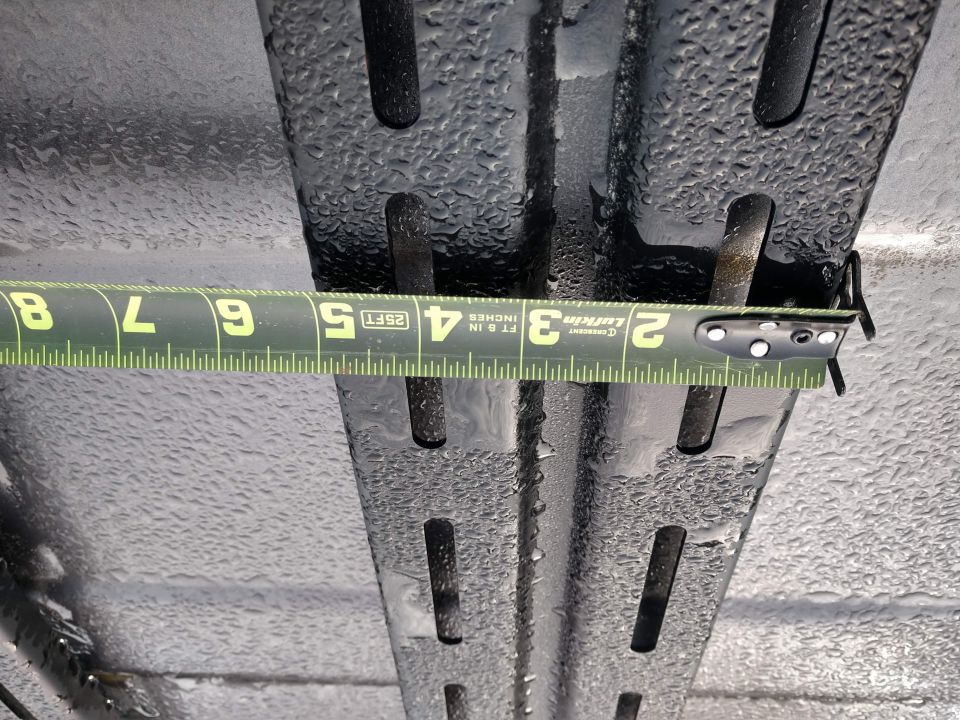 Measuring a black roof rack with a tape measure. The rack has slots and is wet