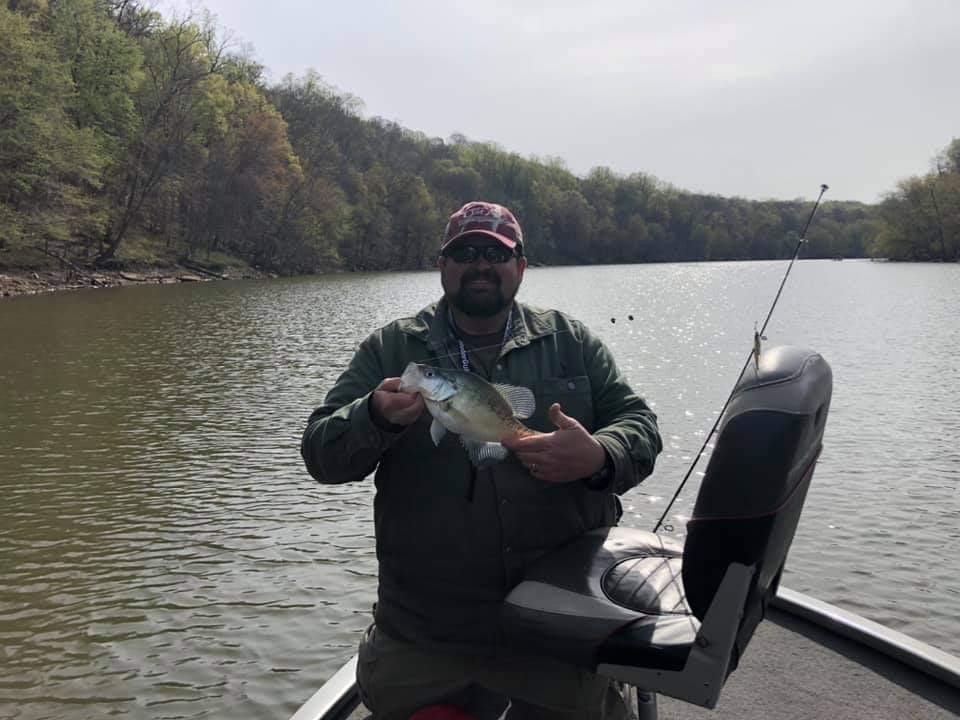 Man holding a crappie on a boat with a fishing rod in the background