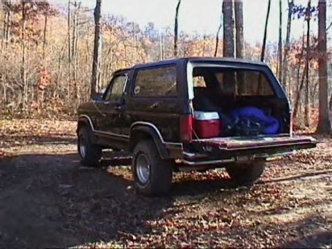1986 Ford F-150 - This black SUV, likely a Ford Bronco from the mid-1980s, is parked outdoors with its tailgate open, revealing camping gear.
