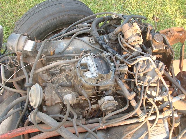 Top view of dirty carbureted V8 engine with intake, alternator and wiring, tire leaning at left