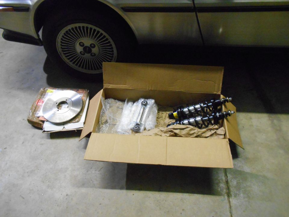 Car parts including shock absorbers and a brake rotor in a box next to a vehicle wheel.