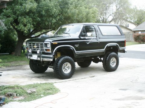 1984 Ford Bronco - This lifted black Ford Bronco is parked on a concrete driveway with trees and houses in the background.