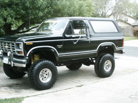 1984 Ford Bronco - This lifted black Ford Bronco is parked on a concrete driveway with trees and houses visible in the background.
