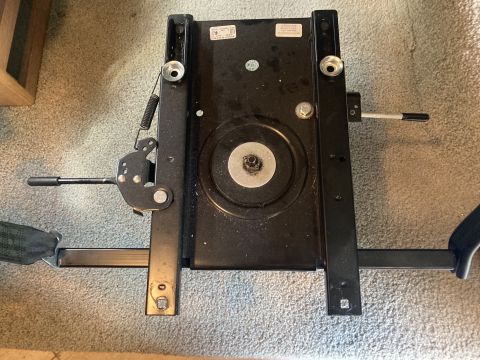 2015 Ford F-550 - A black metal seat swivel base with two adjustment levers, a spring, and a central rotating plate is shown on a carpeted floor.