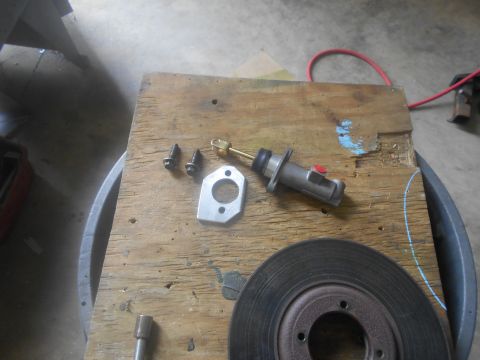 1992 Ford Bronco - A collection of automotive parts, including a brake rotor, a master cylinder, a metal gasket, and bolts, are laid out on a wooden surface.