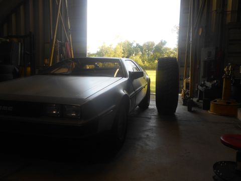1992 Ford Bronco - A silver DeLorean DMC-12 is parked inside a garage with a large tire standing upright next to it. The garage door is open, showing trees an…