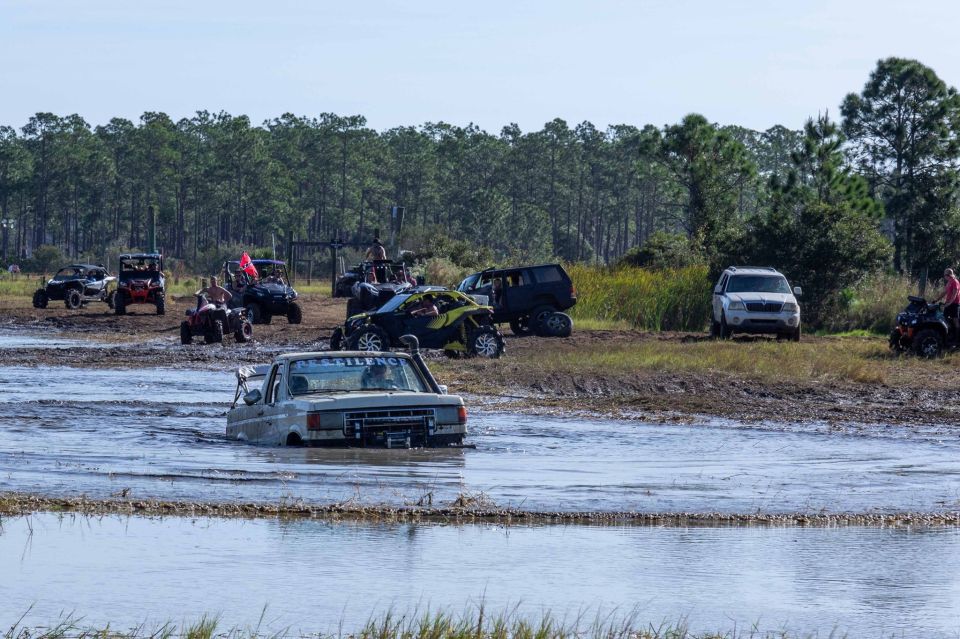 White pickup truck stuck in mud with other off-road vehicles in the background.